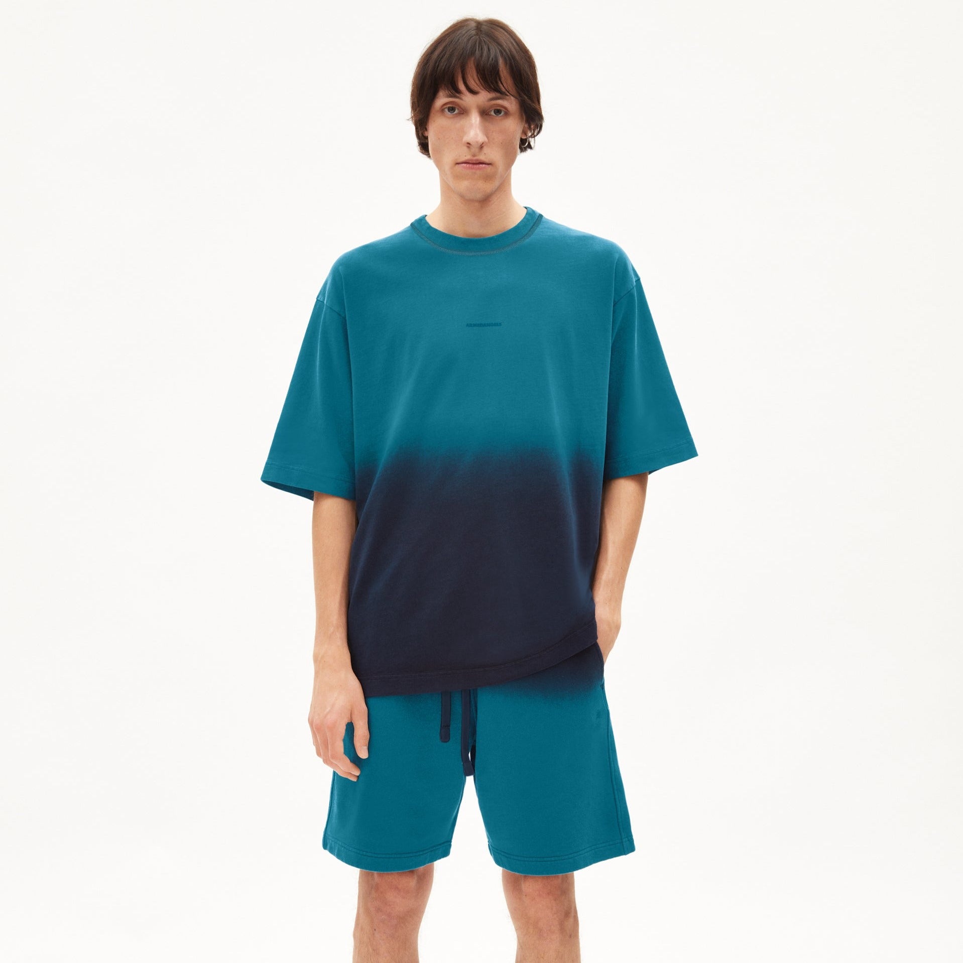 Mann in blau-schwarzem T-Shirt und blauem Shorts steht vor weißem Hintergrund.