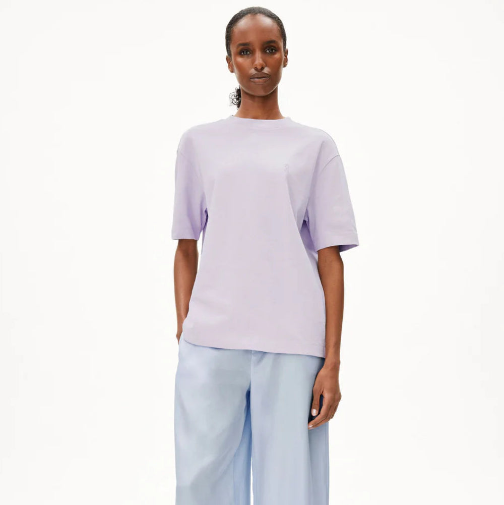 Eine Frau steht vor einem schlichten weißen Hintergrund, trägt ein locker sitzendes lavendelfarbenes T-Shirt, eine hellblaue Hose mit weitem Bein und weiße Schuhe und hat die Hände in den Taschen.