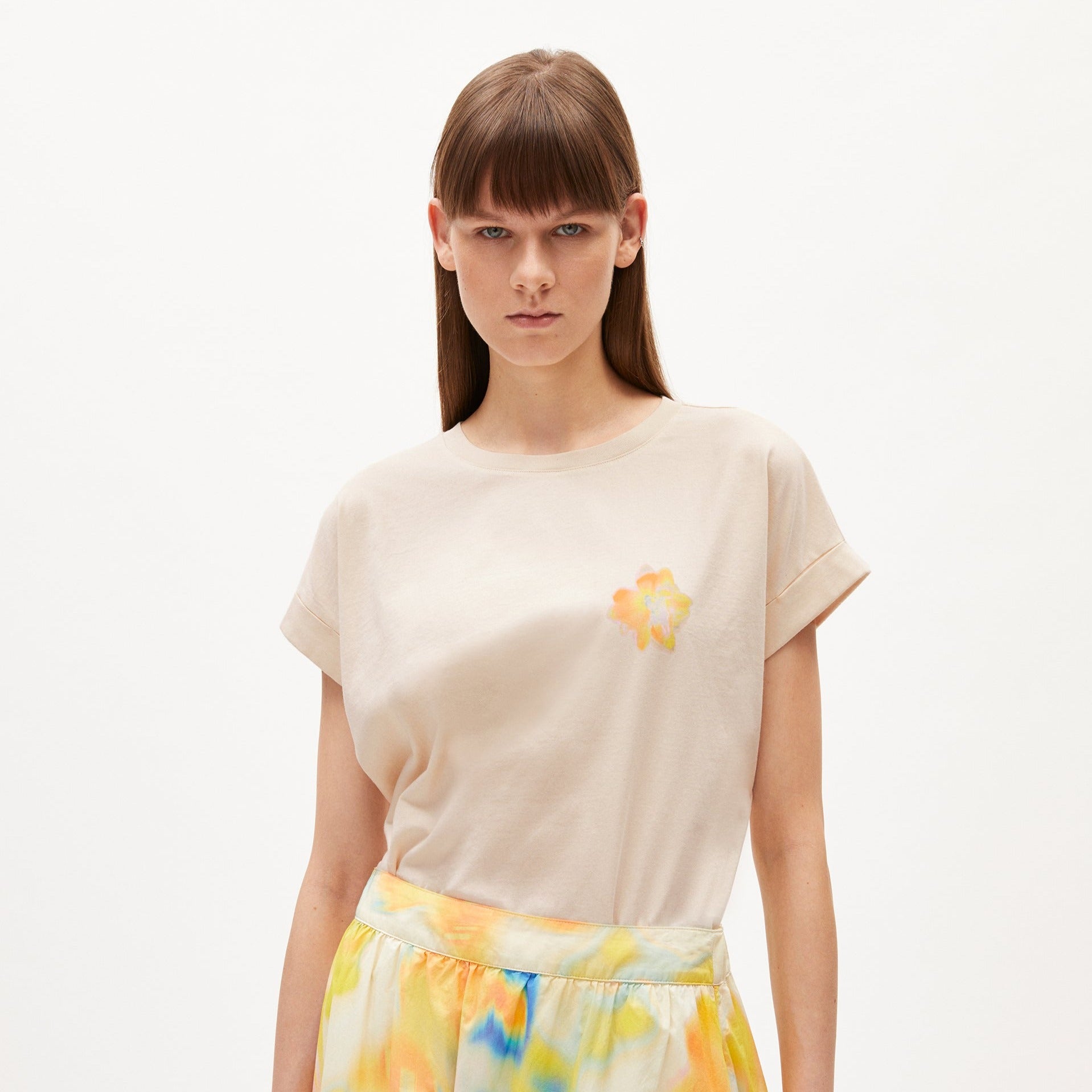 Frau in beigem T-Shirt mit Blumenmuster und einem bunten Rock auf weißem Hintergrund.