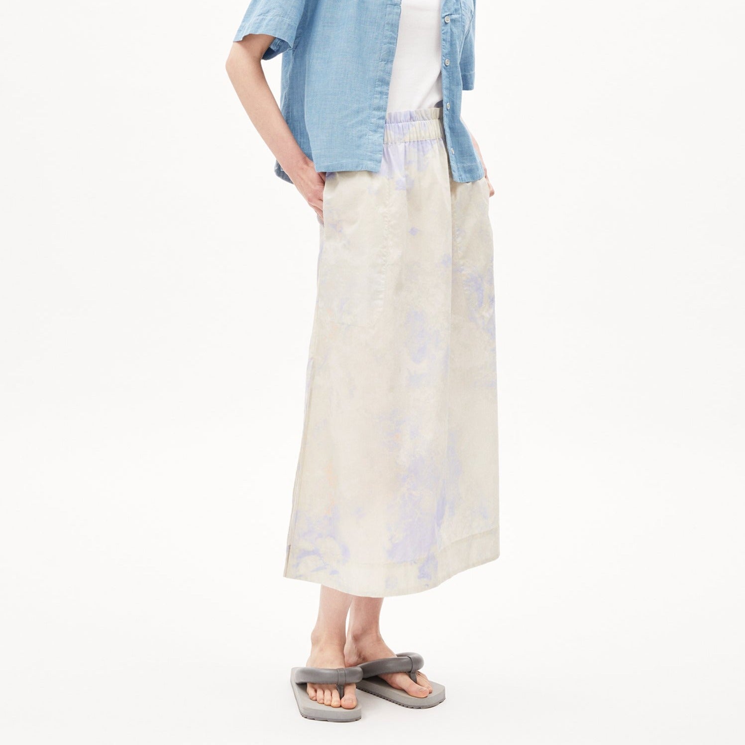 Person in hellblauer Bluse, weißem T-Shirt und langem, beigem Rock, trägt graue Sandalen, vor weißem Hintergrund.