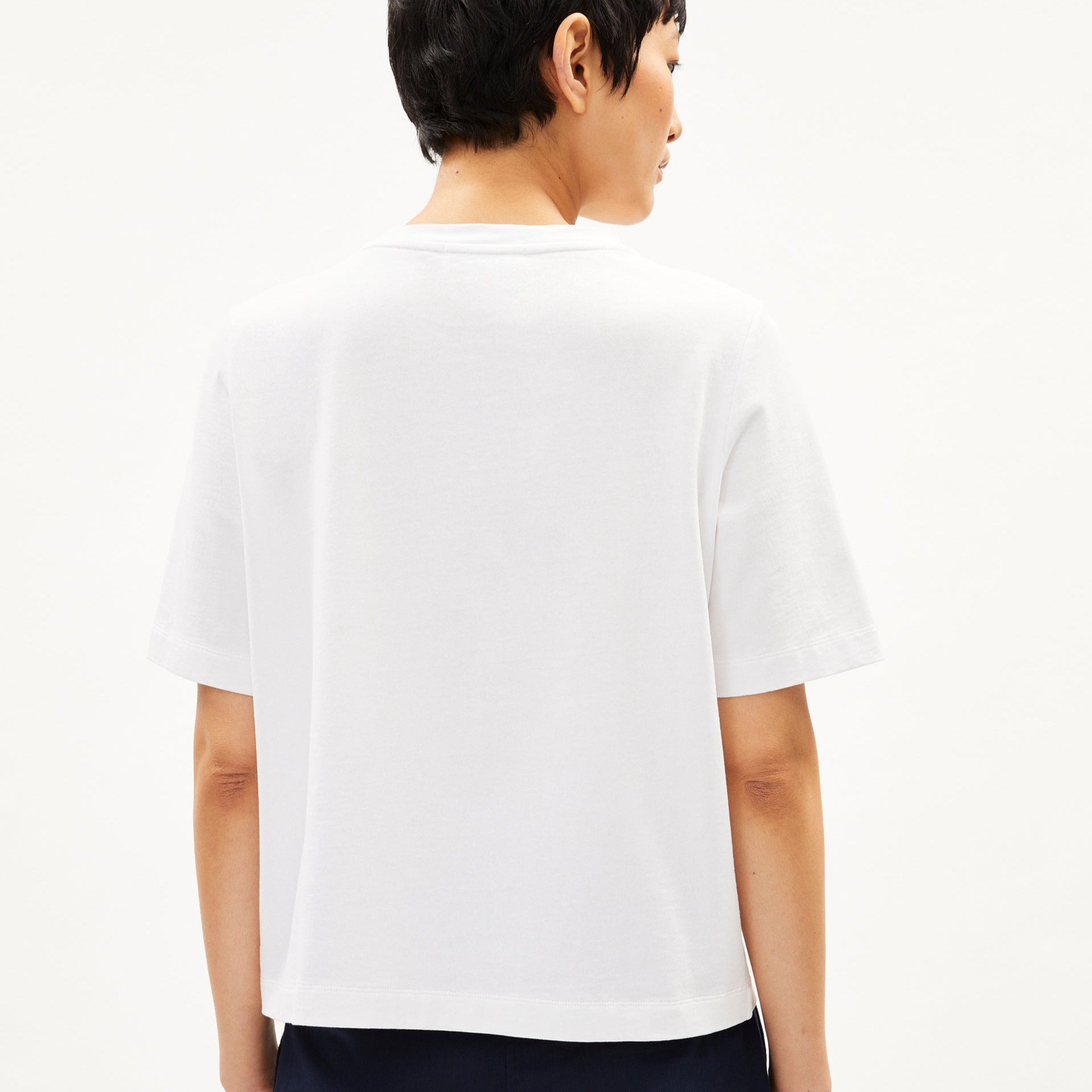 Person von hinten in einem weißen T-Shirt, mit kurzem dunklem Haar, vor einem neutralen Hintergrund.