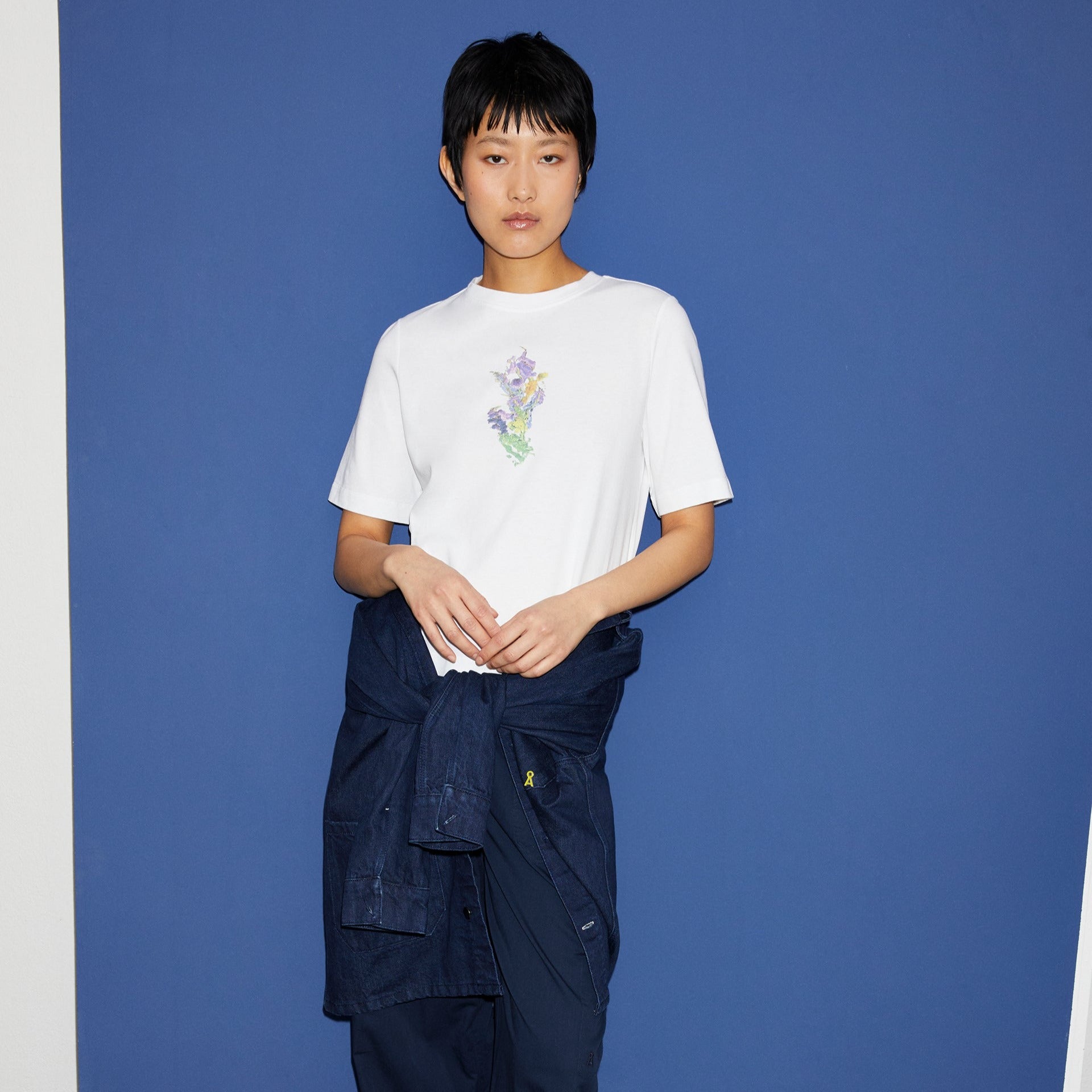 Person in weißem T-Shirt mit floralem Muster und blauer Jacke um die Hüfte steht vor einer dunkelblauen Wand.