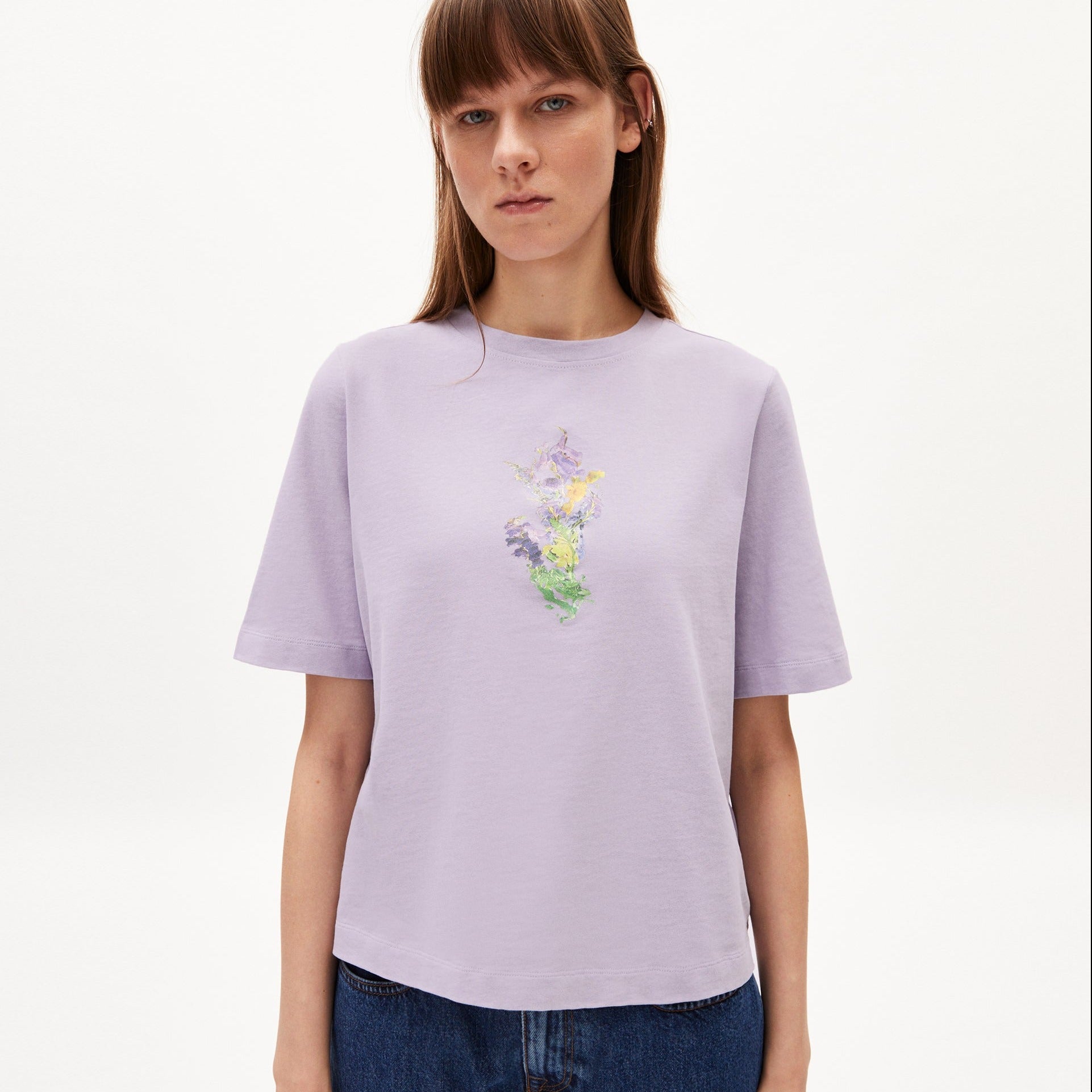 Frau in einem fliederfarbenen T-Shirt mit Blumendruck steht vor einem weißen Hintergrund.