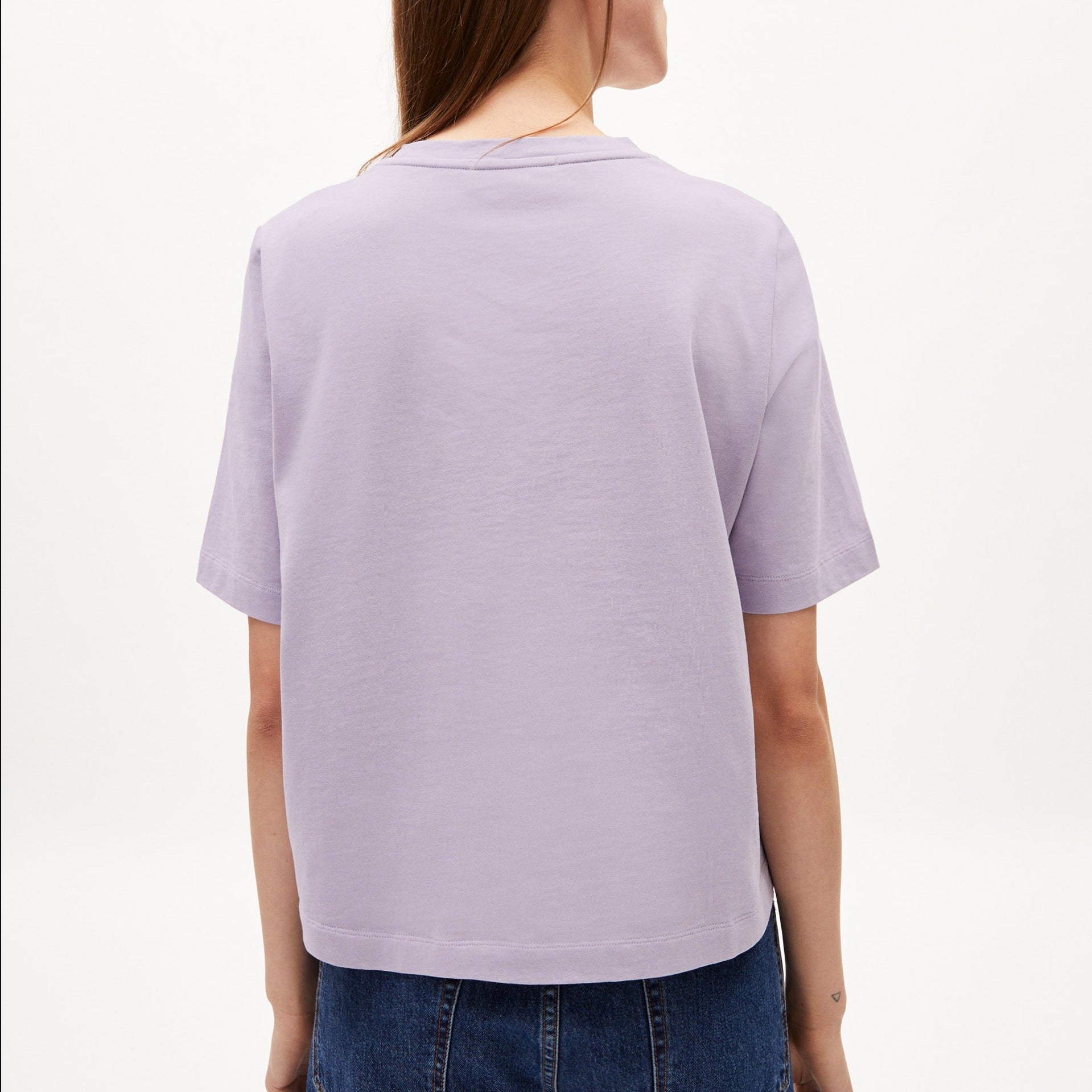 Person in a light purple T-shirt and blue jeans, seen from behind, standing against a plain background.