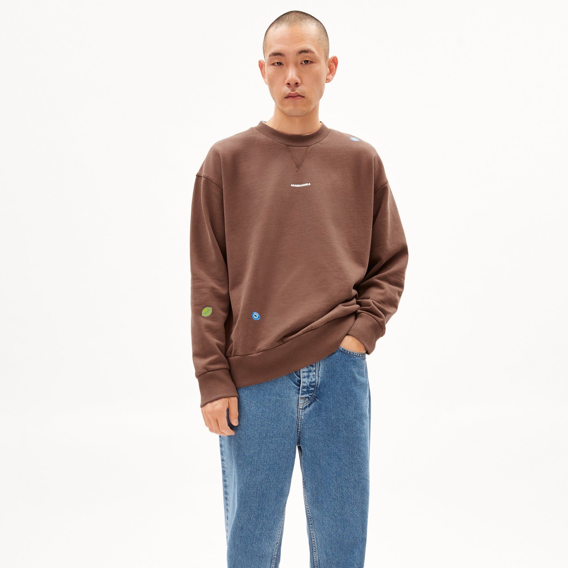 Eine Person in braunem Pullover und blauer Jeans steht vor einem weißen Hintergrund.