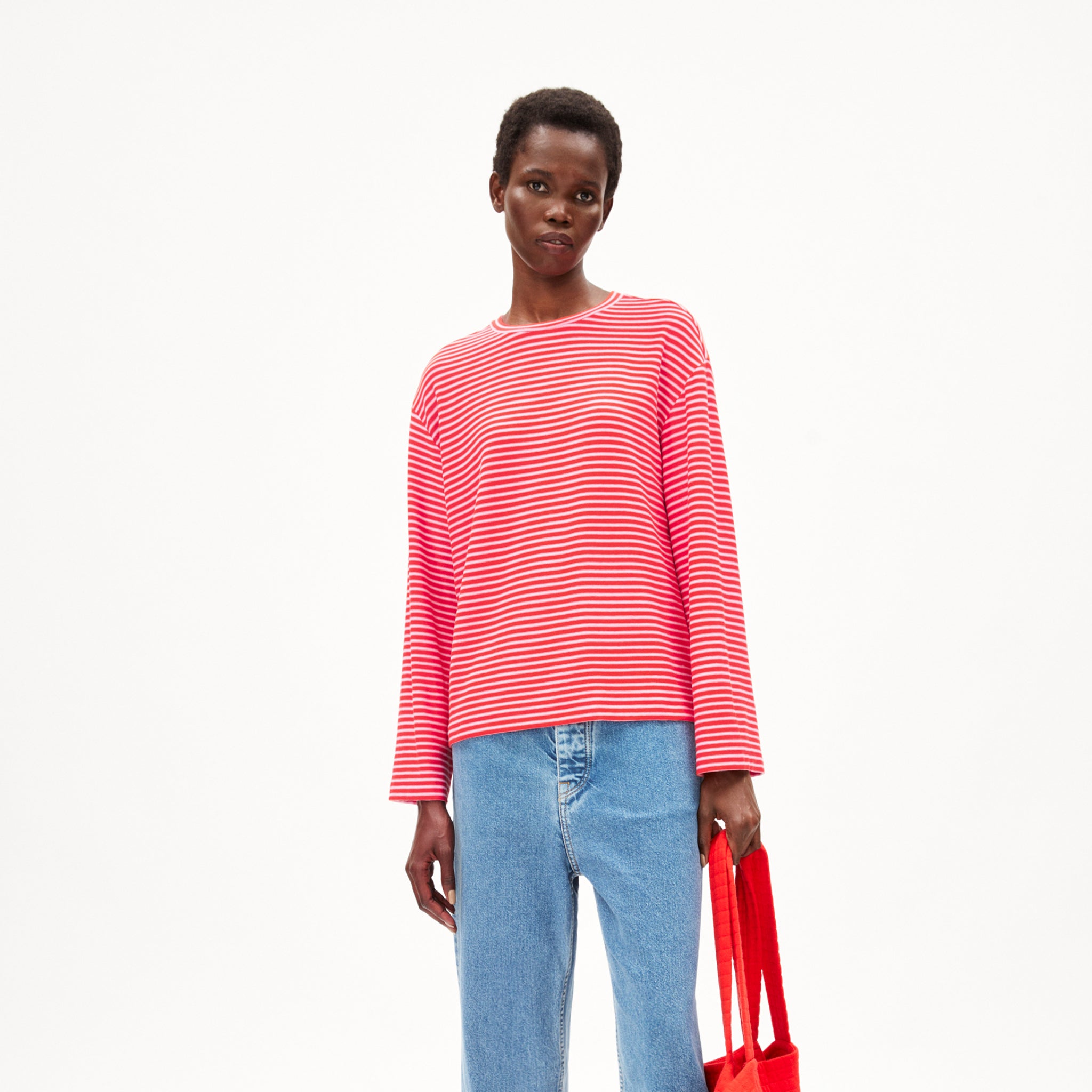 Person in red-orange gestreiftem Pullover und blauer Jeans hält eine rote Tragetasche vor weißem Hintergrund.