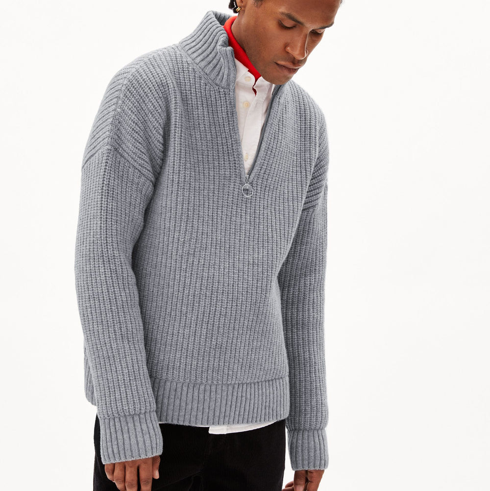 Eine Person, die einen hellgrauen, gerippten Strickpullover über einem weißen Hemd mit Kragen trägt, dessen roter Kragen am Halsausschnitt zu sehen ist, steht vor einem schlichten weißen Hintergrund.