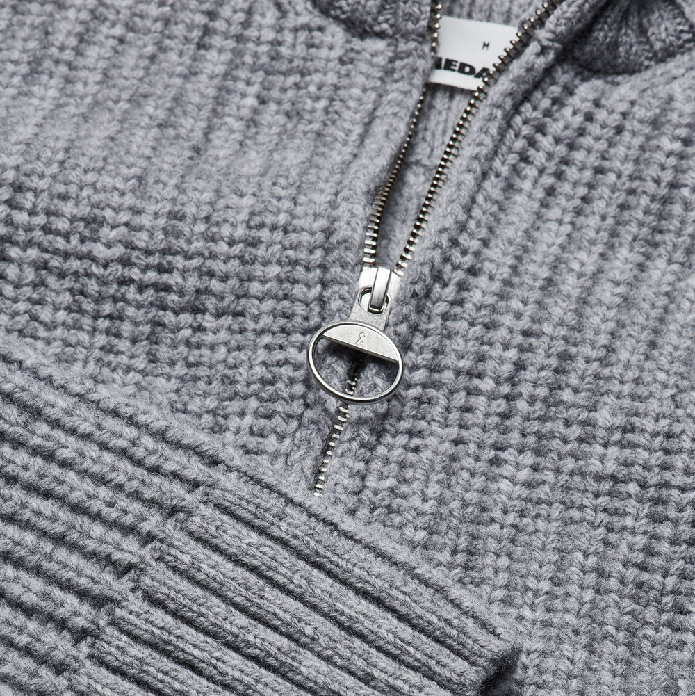 Nahaufnahme eines grauen Strickpullovers mit gerippter Struktur, der teilweise geöffnet wurde, um einen silbernen Reißverschluss und einen kleinen Teil eines weißen Etiketts mit schwarzem Text im Kragen zu zeigen.