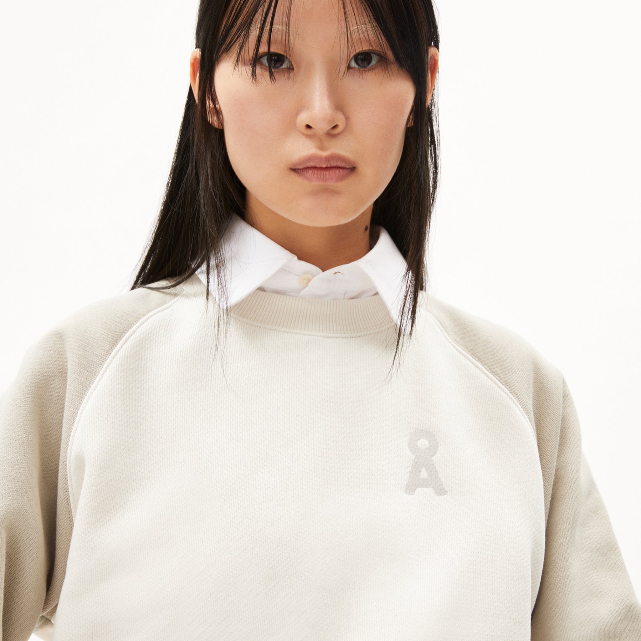 Frau in beigem Pullover mit aufgesticktem Logo, darunter ein weißes Hemd.