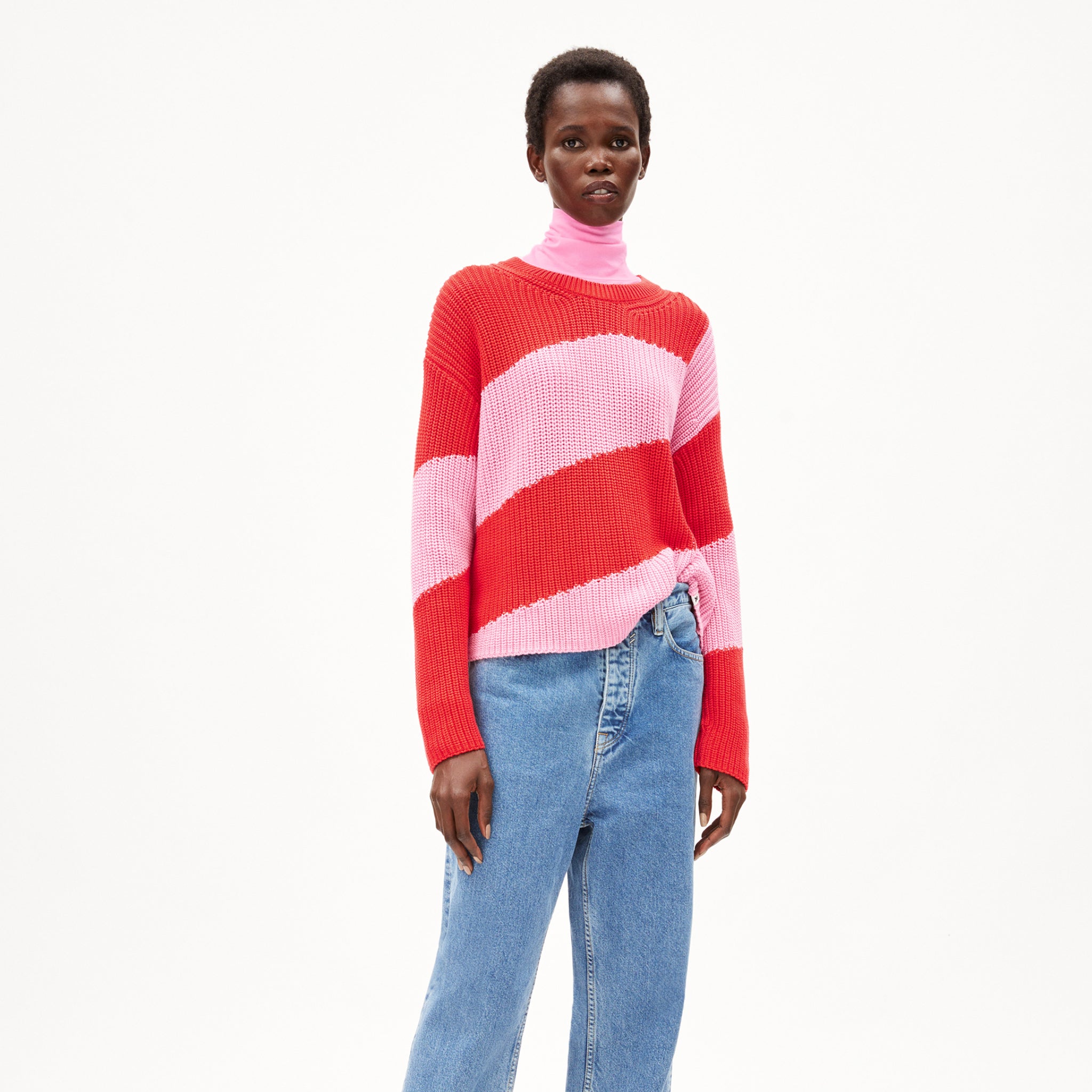 Person in a red and pink striped sweater, pink turtleneck, and blue jeans against a plain white background.