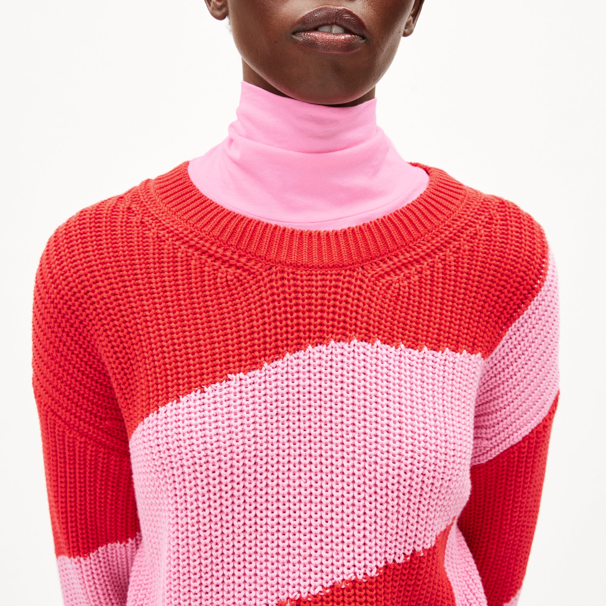 Person in rotem und rosa Pullover und rosa Rollkragenoberteil vor weißem Hintergrund.
