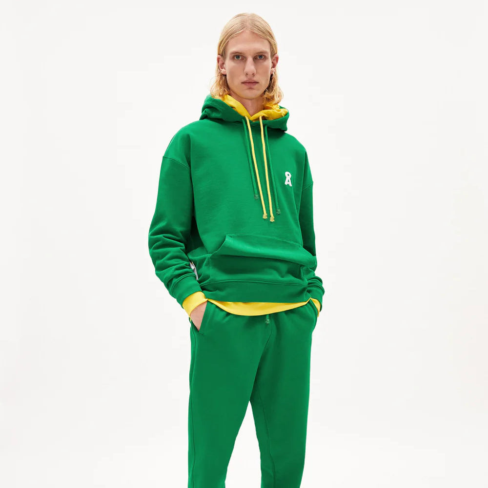 Eine Person mit langen blonden Haaren trägt einen grünen Kapuzenpulli über einem gelben Sweatshirt, eine passende grüne Jogginghose und schwarze Stiefel und steht vor einem schlichten weißen Hintergrund.