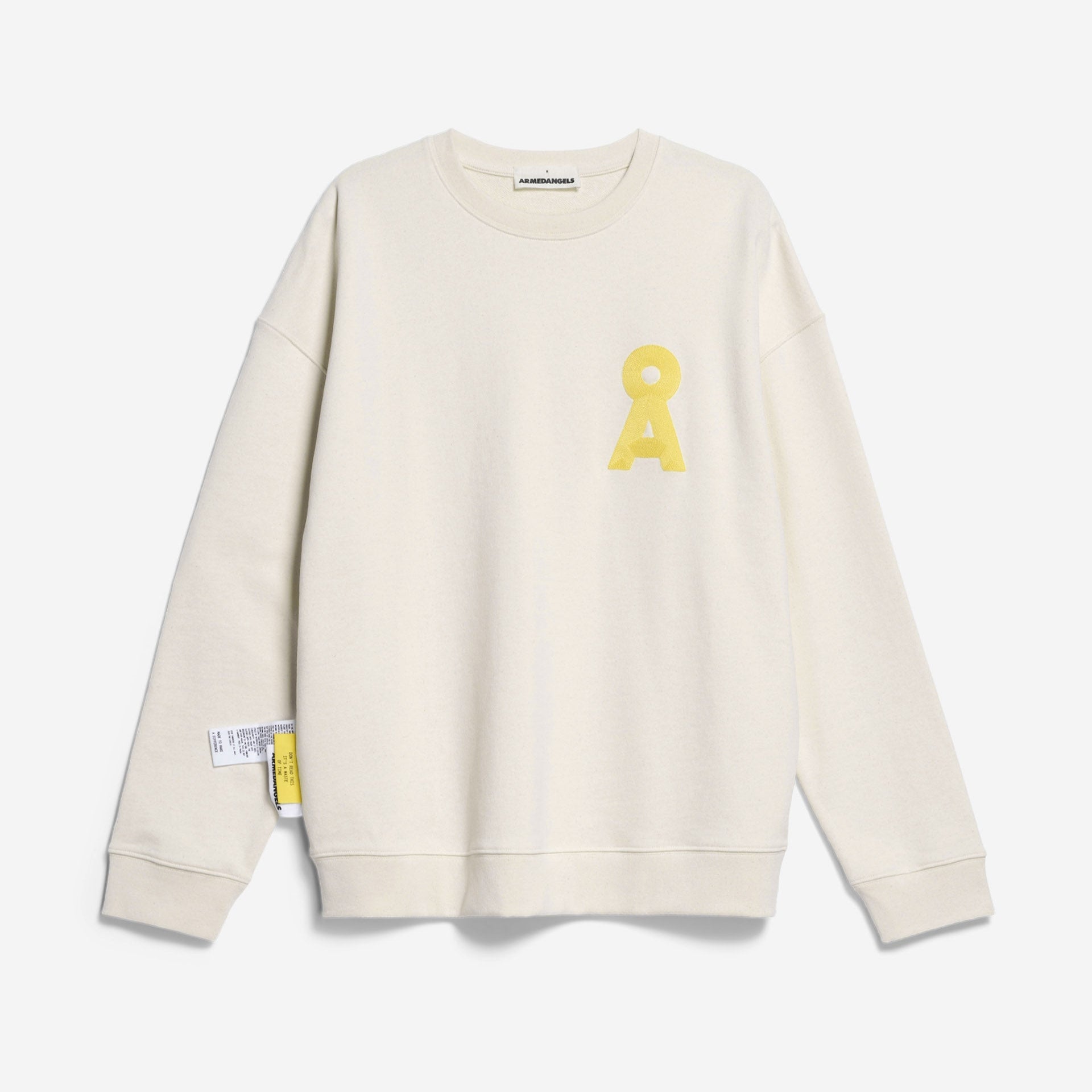 Cremefarbenes Sweatshirt mit langen Ärmeln, mit einem großen gelben A und einem kreisförmigen Symbol auf der linken Brust. Auf der linken Seite in der Nähe des Hosenbundes sind ebenfalls Anhänger angebracht. Der Hintergrund ist einfarbig weiß.