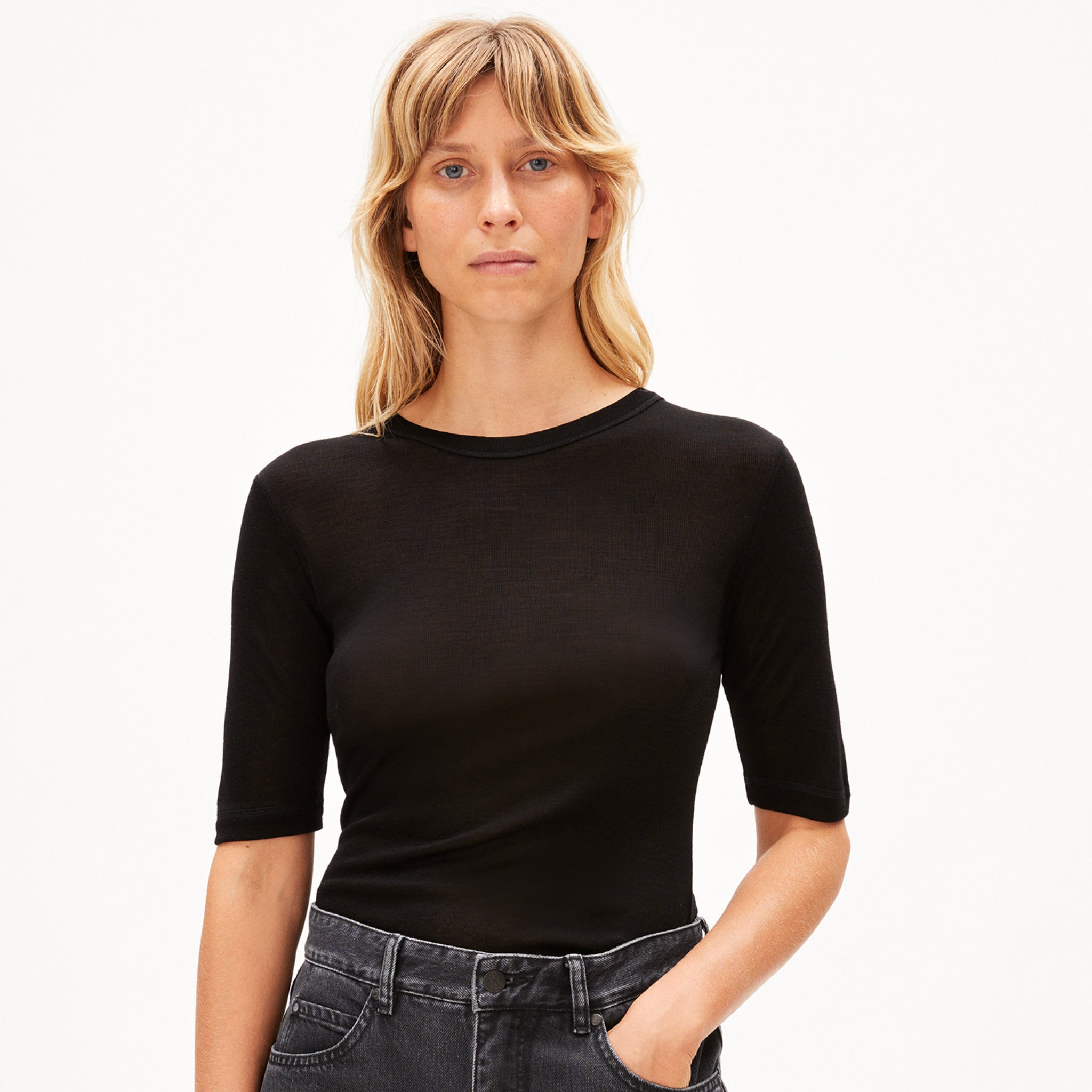 Person mit blondem Haar trägt ein schwarzes T-Shirt und Jeans vor einem weißen Hintergrund.