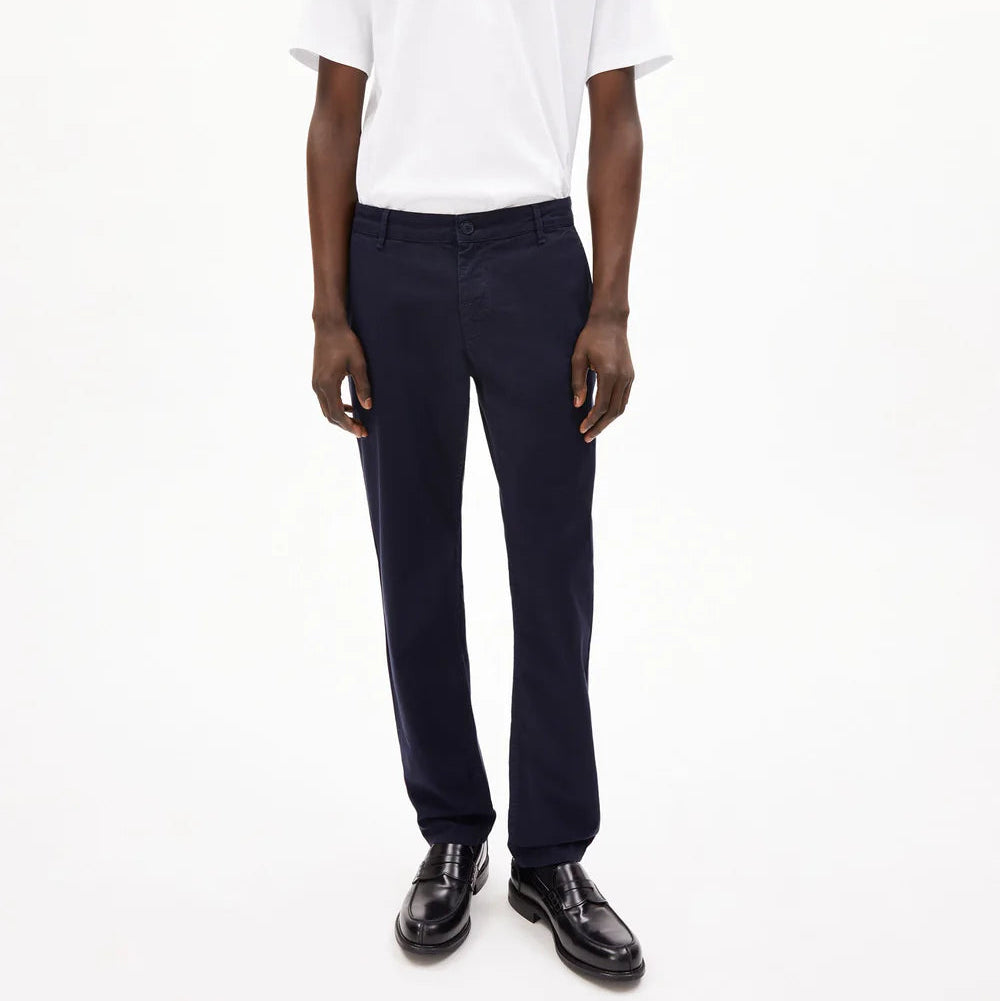 Eine Person steht mit dem Gesicht nach vorne vor einem schlichten weißen Hintergrund und trägt ein weißes kurzärmeliges T-Shirt, eine dunkle marineblaue Hose und schwarze Anzugschuhe.