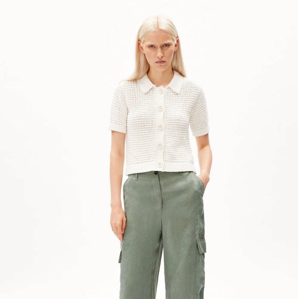 Eine Frau mit langem, glattem blondem Haar trägt ein kurzärmeliges, weißes, geknöpftes Oberteil und eine lockere, salbeigrüne Cargohose. Sie steht mit den Händen in den Taschen vor einem schlichten weißen Hintergrund.