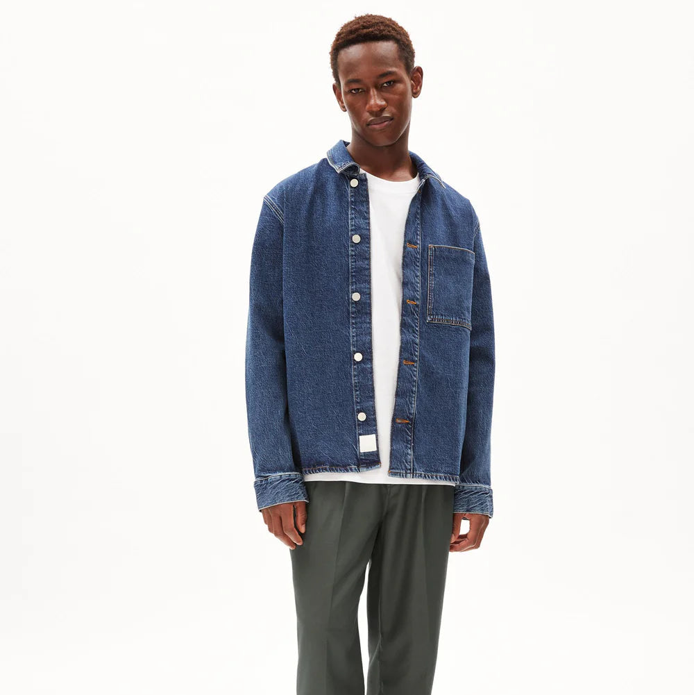 Ein junger Mann steht vor einem schlichten weißen Hintergrund. Er trägt ein blaues Jeanshemd mit Knopfleiste über einem weißen T-Shirt und eine dunkelgrüne Hose. Er schaut mit entspanntem Blick in die Kamera.