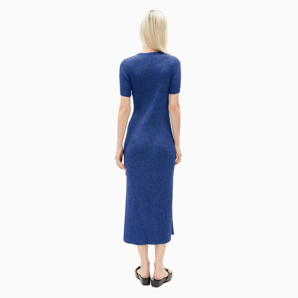 Eine Person mit langem, glattem blondem Haar steht mit dem Gesicht zur Wand und trägt ein kurzärmeliges, blaues, geripptes Midikleid und schwarze Sandalen vor einem schlichten weißen Hintergrund.