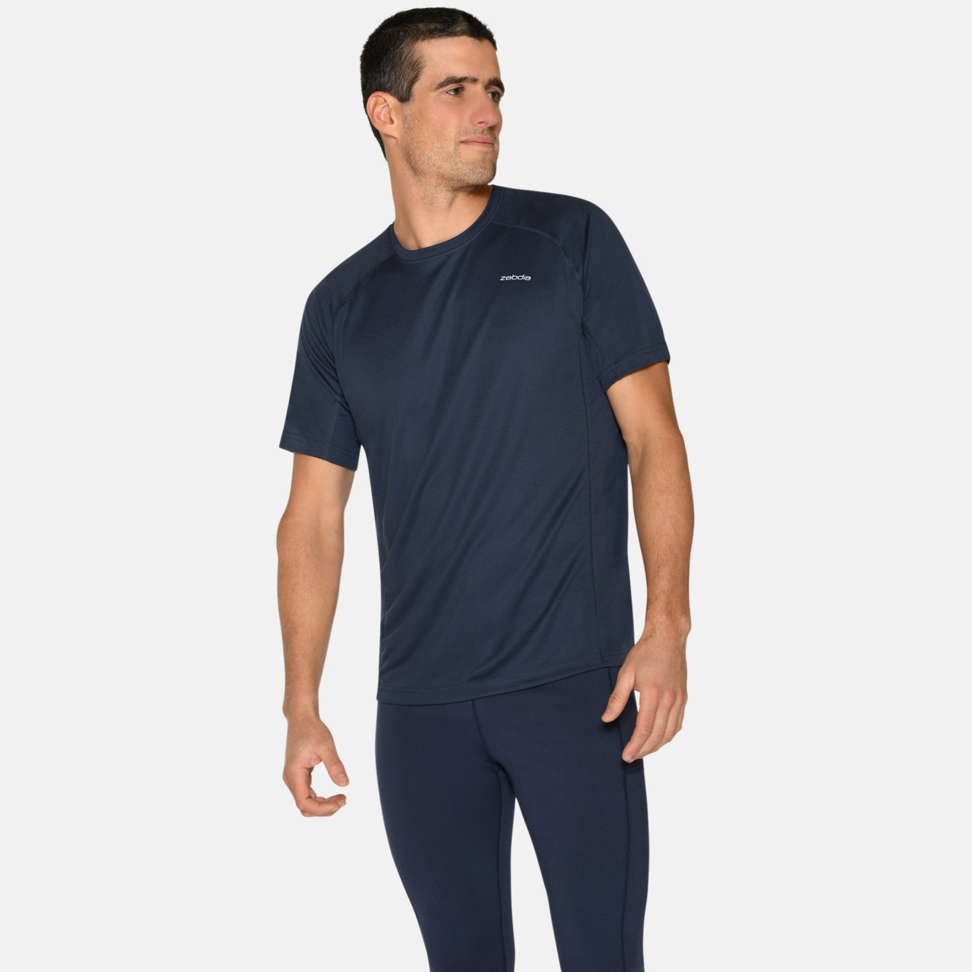 Ein Mann in einem marineblauen Sport-T-Shirt und passenden Leggings steht vor einem schlichten weißen Hintergrund und schaut nach links. Er trägt außerdem blaue Laufschuhe mit orangefarbenen Akzenten.