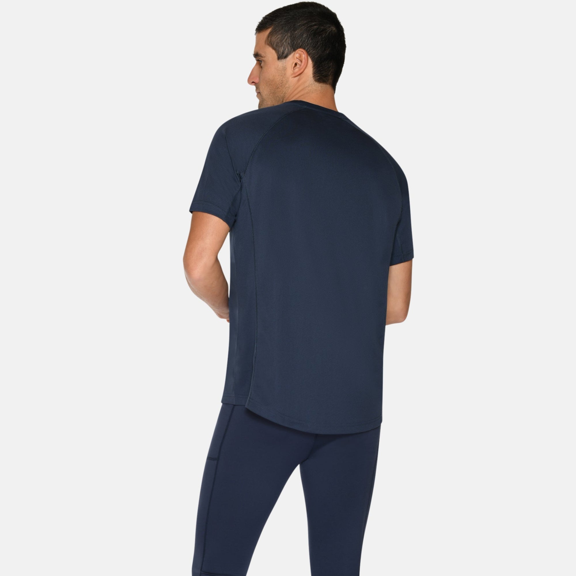 Ein Mann steht mit dem Gesicht zur Wand, trägt ein marineblaues Sport-T-Shirt, marineblaue Laufhosen und blaue Laufschuhe mit gelben Akzenten vor einem schlichten weißen Hintergrund.