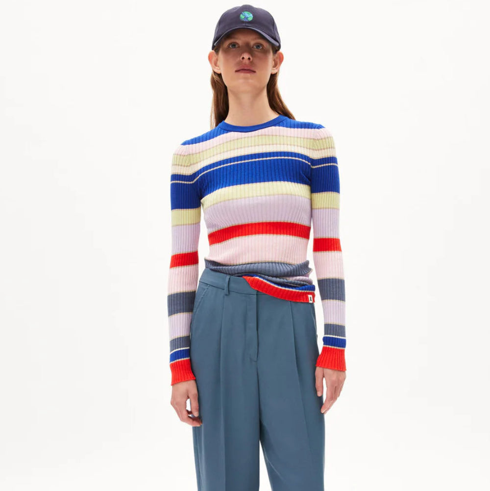 Eine Person, die eine marineblaue Mütze, einen langärmeligen gerippten Pullover mit horizontalen Streifen in Blau, Rot, Gelb und Weiß und eine blaue Hose mit hoher Taille trägt, steht vor einem schlichten weißen Hintergrund.