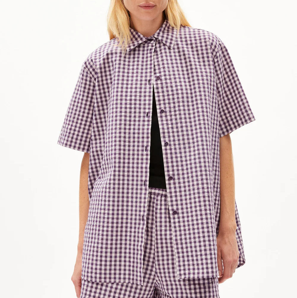 Eine Frau, die ein locker sitzendes, kurzärmeliges, lila-weiß kariertes Button-up-Hemd mit passenden Shorts trägt, steht vor einem schlichten weißen Hintergrund.