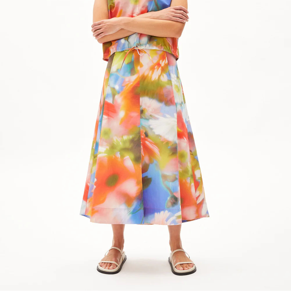 Eine Person steht vor einem weißen Hintergrund und trägt ein ärmelloses, locker sitzendes Oberteil und einen passenden Midirock mit einem lebhaften, bunten Blumenmuster in Orange, Grün, Blau und Weiß. Sie trägt außerdem weiße Sandalen.
