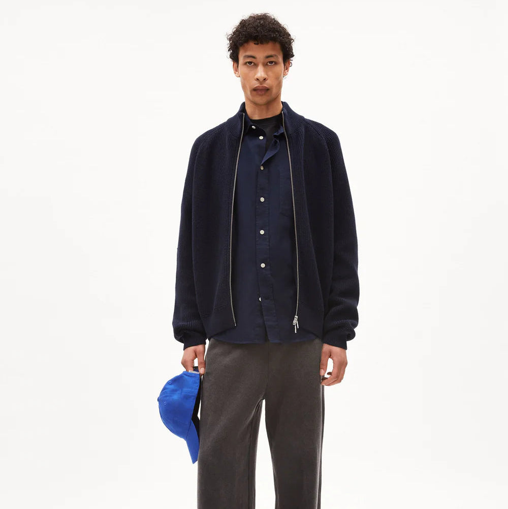 Ein Mann steht vor einem schlichten weißen Hintergrund, trägt eine marineblaue Jacke mit Reißverschluss über einem marineblauen Hemd, eine weite dunkelgraue Hose und schwarze Schuhe und hält eine hellblaue Baseballmütze in der linken Hand.