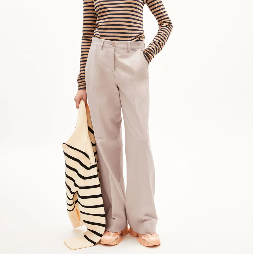 Eine Frau steht vor einem weißen Hintergrund, trägt einen braun-schwarz gestreiften Rollkragenpullover, eine hellbeige Hose mit weitem Bein und pfirsichfarbene Schuhe und hat einen gestreiften Pullover über den linken Arm gezogen.