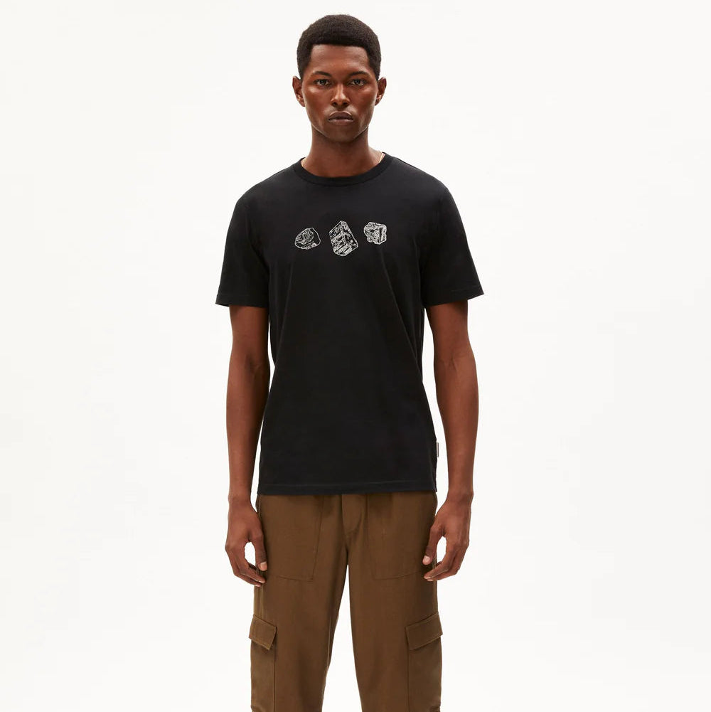 Ein Mann steht mit dem Gesicht nach vorne vor einem weißen Hintergrund. Er trägt ein schwarzes T-Shirt mit drei abgebildeten Fäusten auf der Brust, eine braune Cargohose und weinrote Turnschuhe.