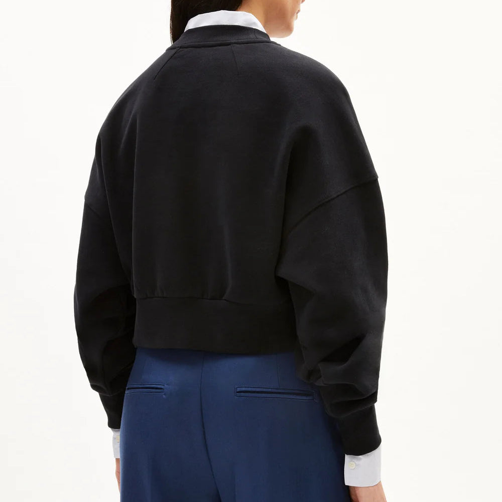 Eine Person, die mit dem Rücken zur Kamera steht und einen schwarzen Kurzpullover über einem weißen Hemd mit Kragen und eine blaue Hose vor einem schlichten weißen Hintergrund trägt.