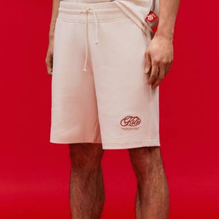 Eine Person, die cremefarbene Shorts mit einem KitKat-Logo, ein cremefarbenes Hemd, bordeauxfarbene Socken und cremefarbene Slipper-Sandalen trägt, steht vor einem leuchtend roten Hintergrund. Nur die untere Hälfte des Körpers ist sichtbar.