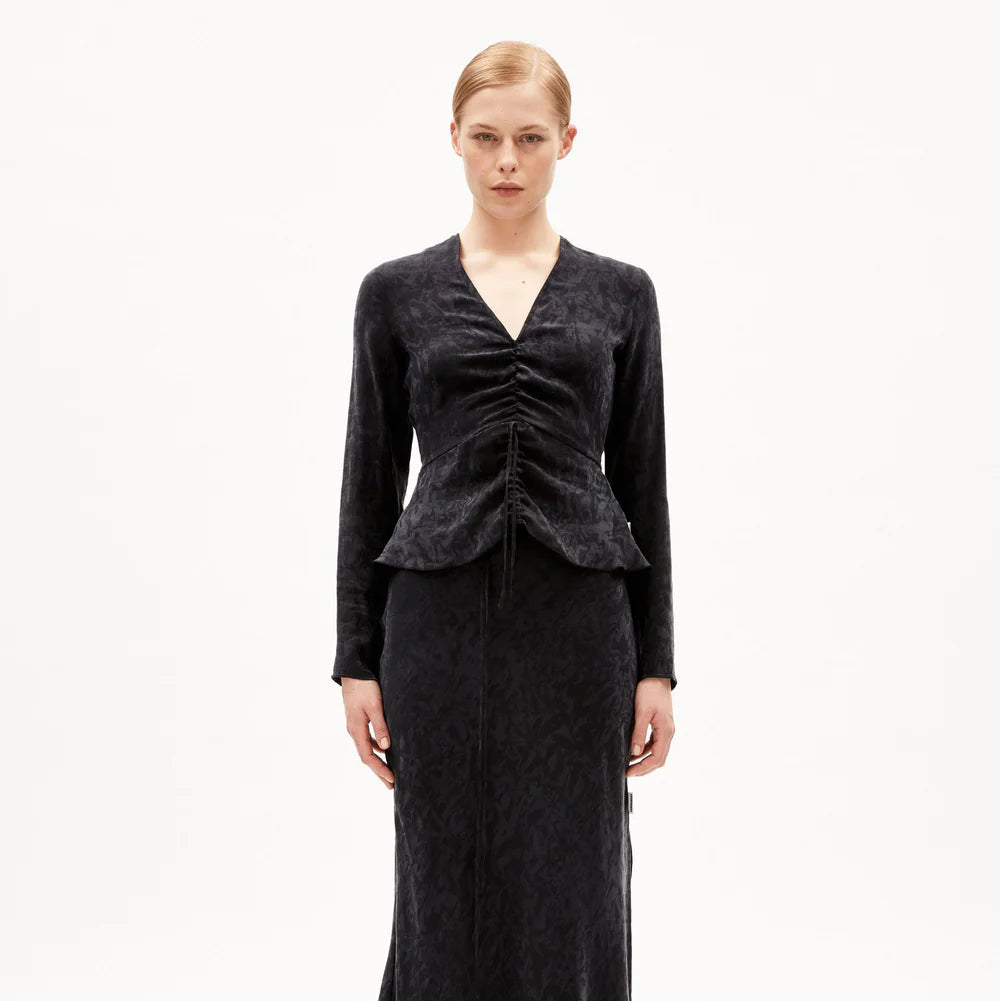 Eine Frau mit zurückgebundenen blonden Haaren trägt ein langärmeliges, tailliertes schwarzes Kleid mit V-Ausschnitt und Rüschen an der Taille vor einem schlichten weißen Hintergrund.