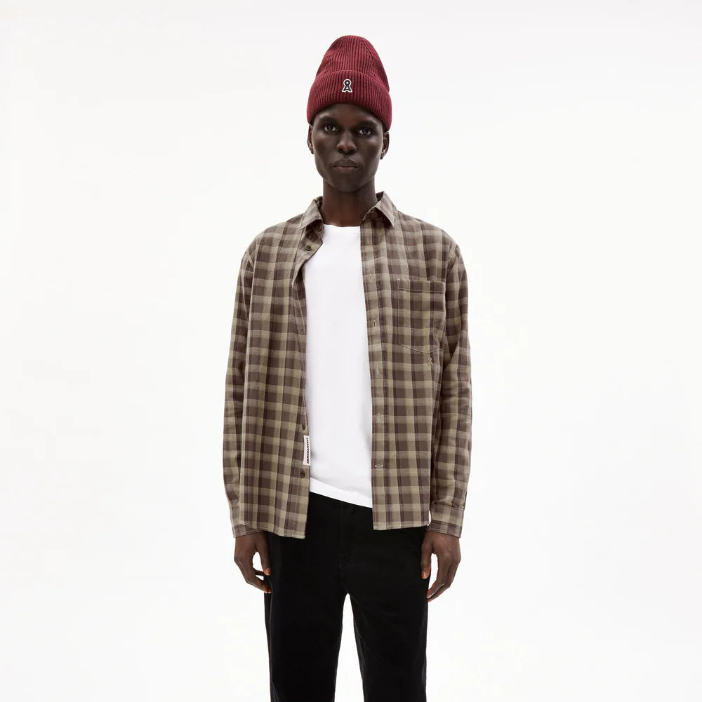 Eine Person, die eine rote Strickmütze, ein braun kariertes Button-up-Hemd über einem weißen T-Shirt und eine schwarze Hose trägt, steht vor einem schlichten weißen Hintergrund.