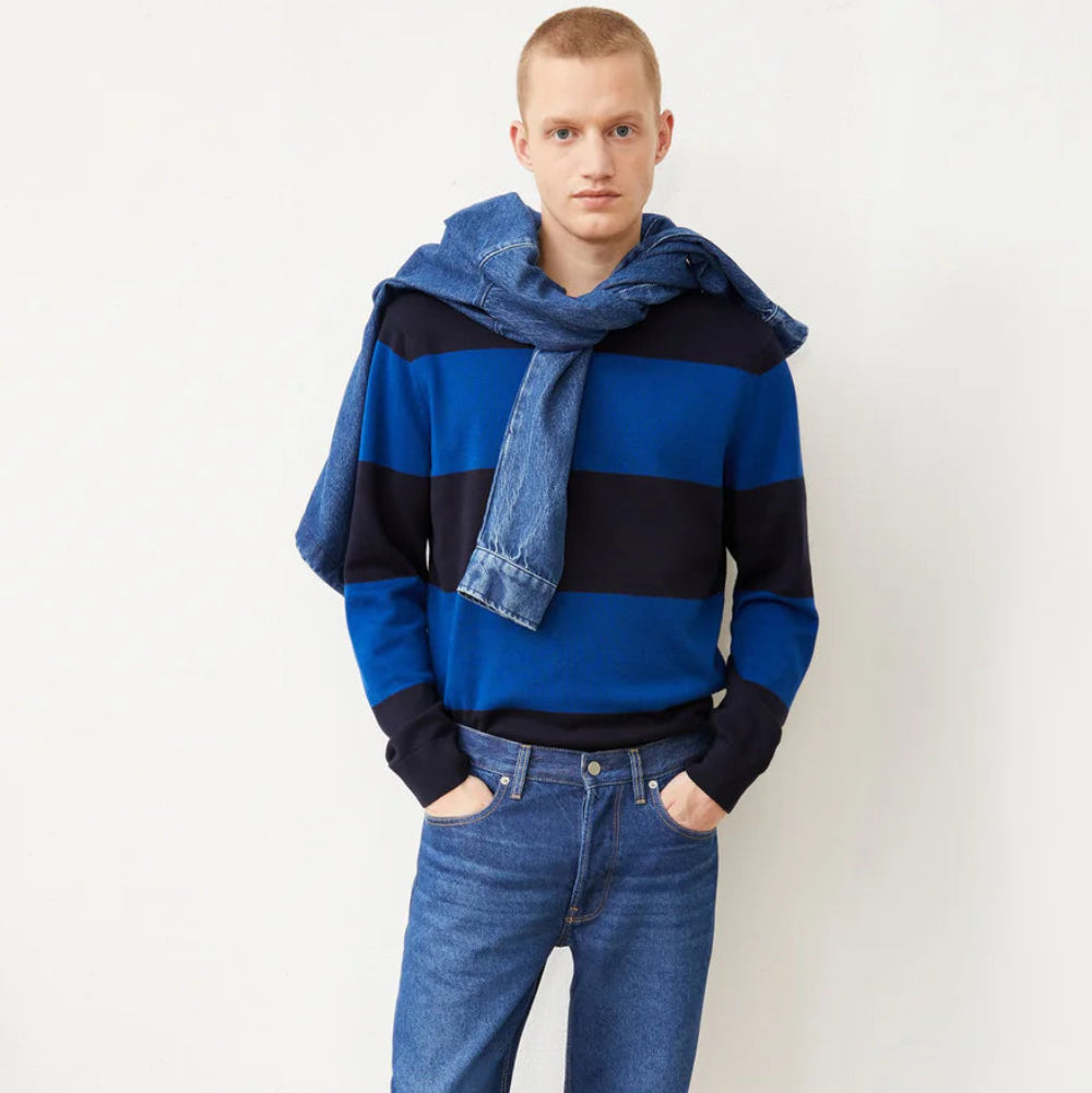 Ein Mann steht vor einer schlichten weißen Wand und trägt einen blau-schwarz gestreiften Pullover, blaue Jeans, schwarze Schuhe und eine über die Schultern gezogene Jeansjacke. Er hat kurze Haare und einen neutralen Gesichtsausdruck.