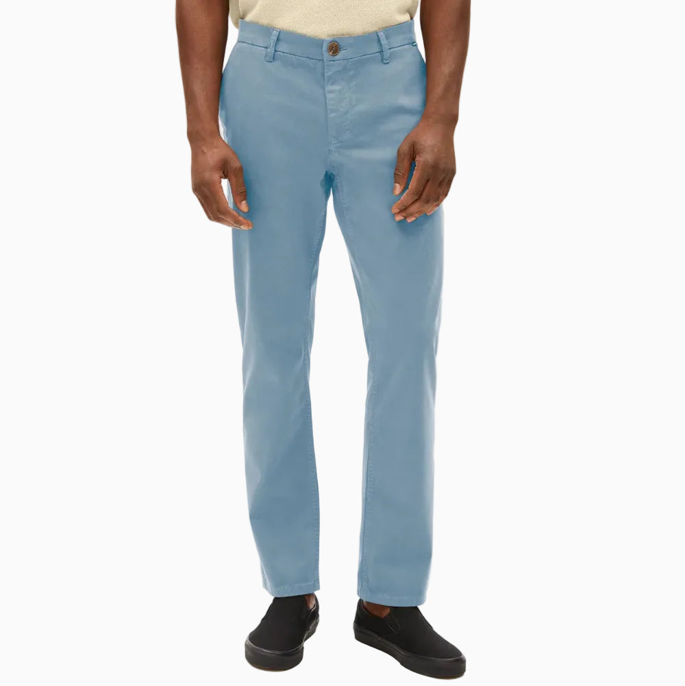 Eine Person, die eine hellblaue Chinohose, ein hellbeiges kurzärmeliges Hemd und schwarze Slipper trägt, steht vor einem schlichten weißen Hintergrund.