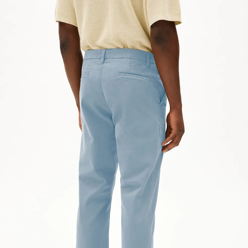 Eine Person, die eine hellblaue Hose, ein beiges kurzärmeliges Hemd und schwarze Slipper trägt, steht mit dem Rücken zur Kamera vor einem schlichten weißen Hintergrund.