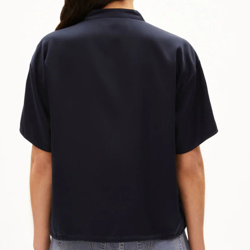 Eine Person mit langen, dunklen Haaren ist von hinten zu sehen. Sie trägt ein dunkles, kurzärmeliges Hemd und hellblaue Jeans vor einem schlichten weißen Hintergrund.