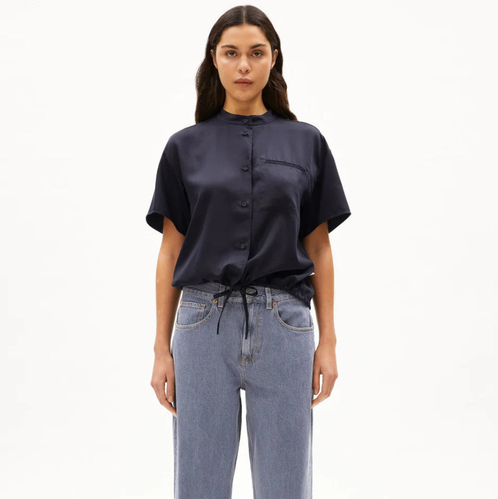 Eine Person steht mit dem Gesicht nach vorne und trägt ein kurzärmeliges, marineblaues, geknöpftes Hemd mit Kordelzug in der Taille, locker sitzende hellblaue Jeans und beigefarbene Schuhe vor einem schlichten weißen Hintergrund.