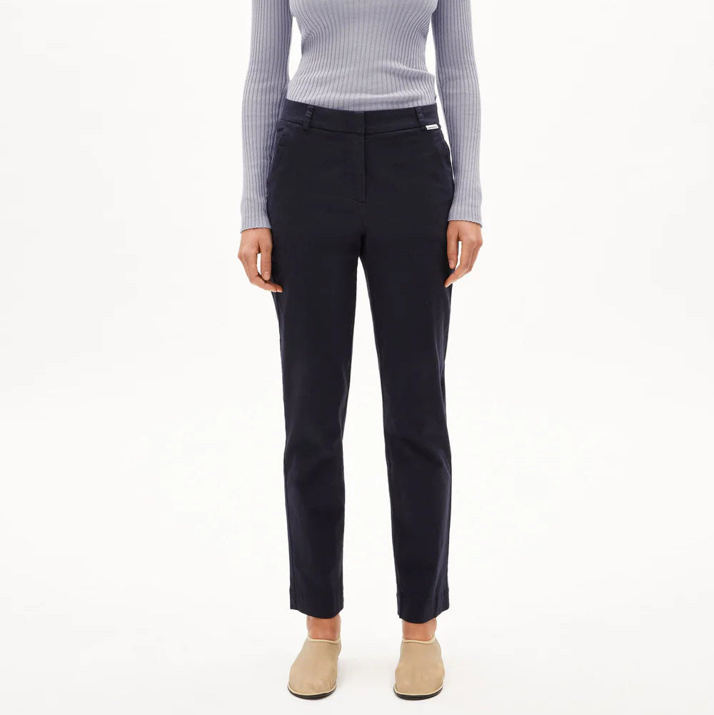 Eine Frau steht mit dem Gesicht nach vorne, trägt ein hellgraues, geripptes, langärmeliges Oberteil, eine marineblaue Hose und beigefarbene Slipper vor einem schlichten weißen Hintergrund.