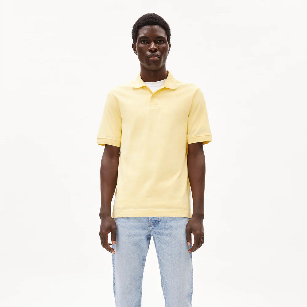 Ein Mann, der ein hellgelbes, kurzärmeliges Poloshirt und hellblaue Jeans trägt, steht mit dem Gesicht nach vorne vor einem schlichten weißen Hintergrund.