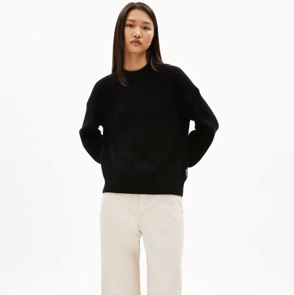 Eine Person mit glattem, langem, dunklem Haar trägt einen schwarzen Pullover, eine cremefarbene Hose mit weitem Bein und schwarze Schuhe und steht vor einem schlichten weißen Hintergrund.