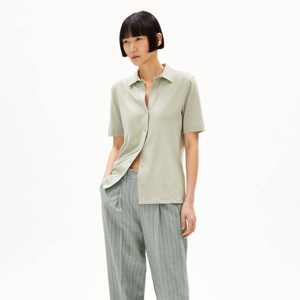 Eine Person mit kurzen schwarzen Haaren trägt ein hellgrünes, kurzärmeliges, geknöpftes Hemd, eine lockere, hellgraue Nadelstreifenhose, die an den Knöcheln zusammengeschnürt ist, und grüne Sandalen. Sie steht mit einer Hand in der Tasche vor einem weißen Hintergrund.