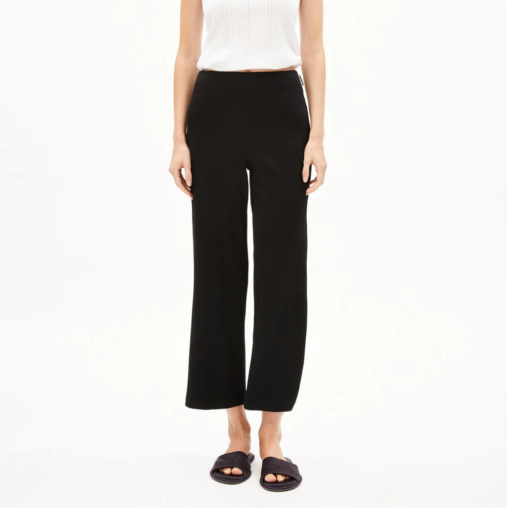 Eine Frau steht mit dem Gesicht nach vorne, trägt ein weißes ärmelloses Oberteil, eine schwarze Hose mit hoher Taille und schwarze Slide-Sandalen vor einem schlichten weißen Hintergrund.