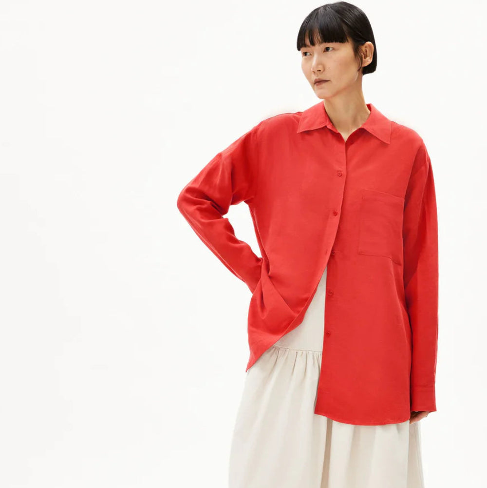 Eine Person steht vor einem schlichten weißen Hintergrund, trägt ein leuchtend rotes, langärmeliges Hemd über einem fließenden weißen Rock und schwarze Sandalen. Sie hat glattes dunkles Haar mit Pony und posiert mit einer Hand in der Hüfte.