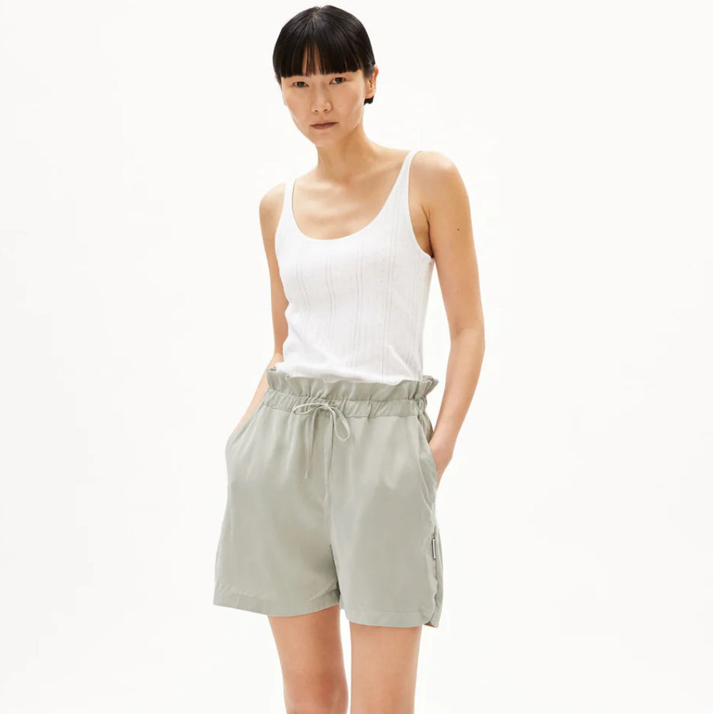 Eine Person steht mit den Händen in den Taschen einer hellgrauen Kordelhose, trägt ein weißes ärmelloses Oberteil und schwarze Sandalen mit grünen Akzenten vor einem schlichten weißen Hintergrund.