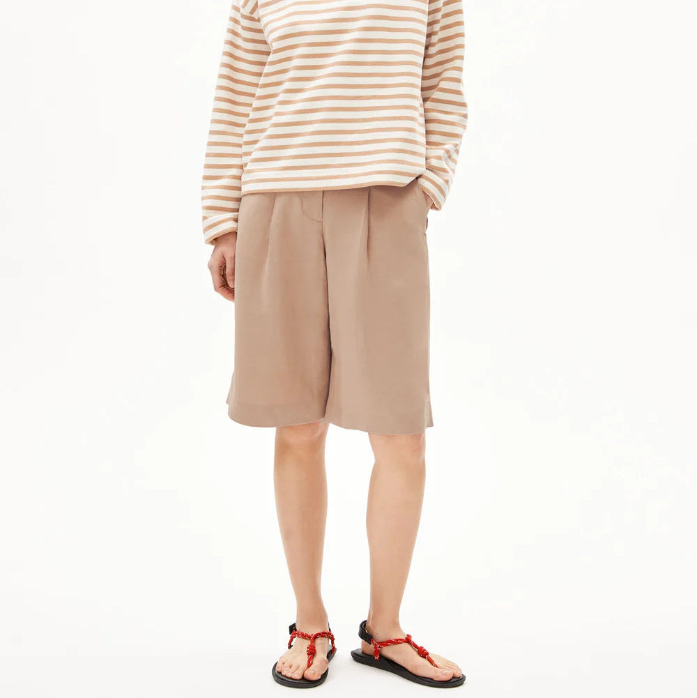 Eine Frau mit kurzen schwarzen Haaren trägt ein beige-weiß gestreiftes, langärmeliges Hemd, weite beigefarbene, knielange Shorts und rot-schwarze Sandalen und steht vor einem schlichten weißen Hintergrund.