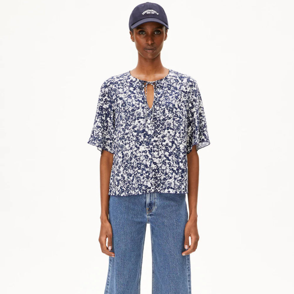 Eine Person, die eine marineblaue Baseballkappe, eine blau-weiß geblümte kurzärmelige Bluse mit Schlüssellochausschnitt, eine weit geschnittene Bluejeans und schwarze Slide-Sandalen trägt, steht vor einem schlichten weißen Hintergrund.