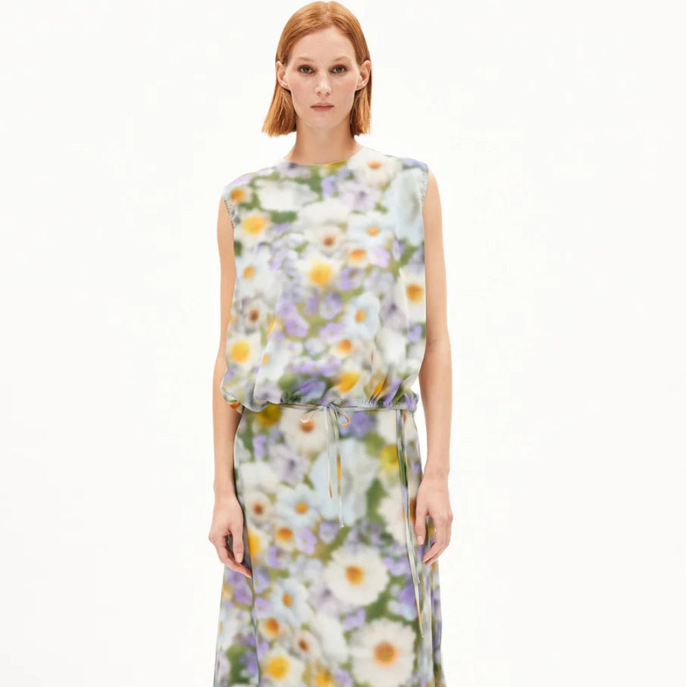 Eine Person mit glattem, schulterlangem Haar trägt ein ärmelloses, langes, lockeres Kleid mit einem bunten, verschwommenen Blumenmuster, das in der Taille mit einer dünnen Krawatte zusammengehalten wird, und kombiniert es mit schwarzen Sandalen vor einem schlichten weißen Hintergrund.