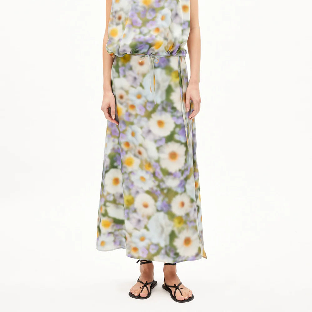 Eine Frau mit glattem, schulterlangem Haar trägt ein ärmelloses, langes Kleid mit einem verschwommenen weißen und gelben Gänseblümchenmuster, das in der Taille mit einem dünnen Gürtel gerafft ist. Sie steht barfuß auf einem schlichten weißen Hintergrund.